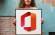 Eine junge Frau steht vor einer etwas verwitterten Mauer; in ihren Händen hält sie ein Bild mit dem Logo von Microsoft Office