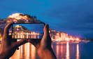 Ein Smartphone in den Händen einer Person, die damit gerade eine Abendaufnahme einer Insel-Ortschaft macht